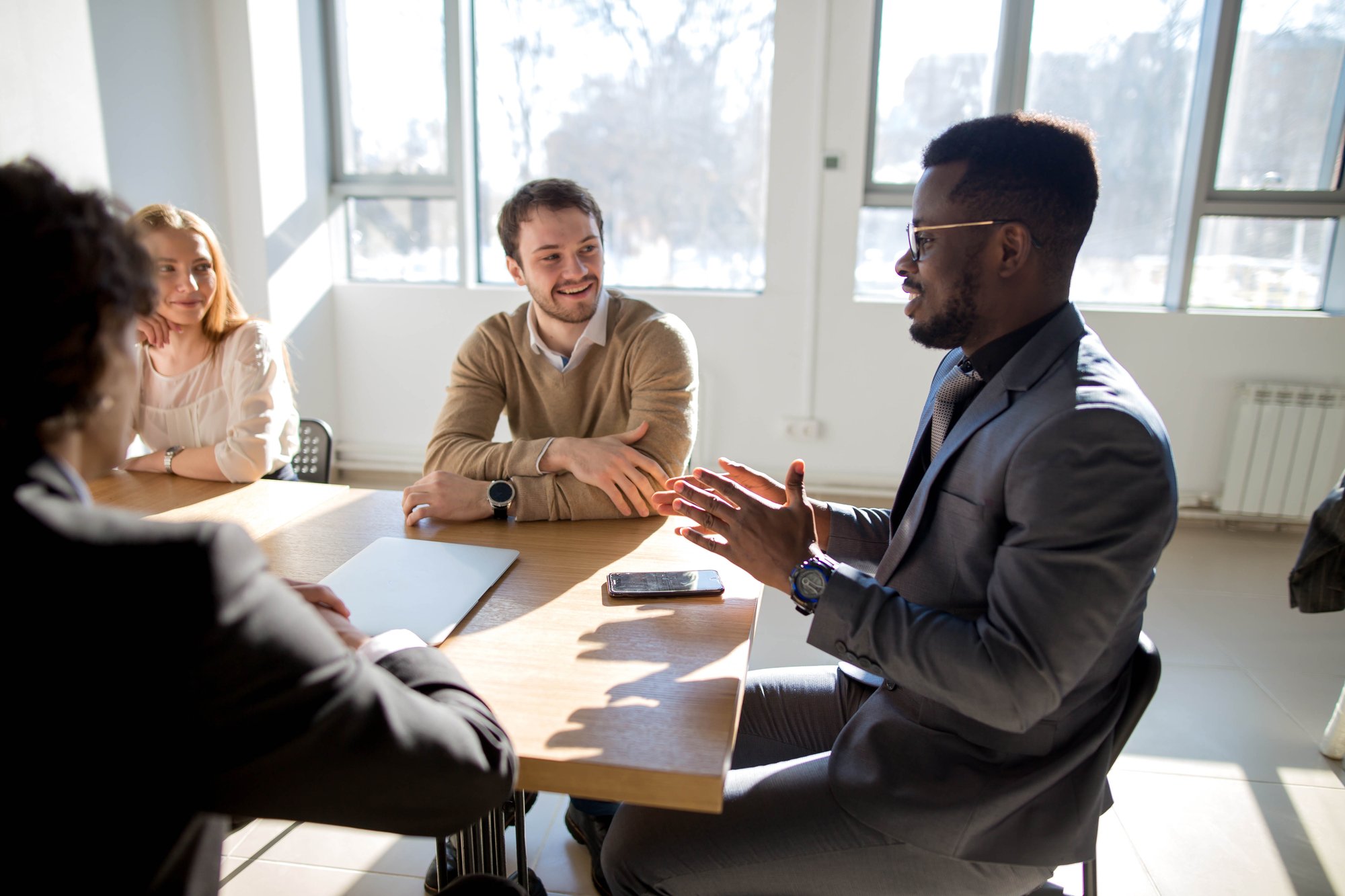 Four people seated around a table in a bright room, engaged in conversation. One person gestures with hands while speaking. A smartphone and notebook are on the table.